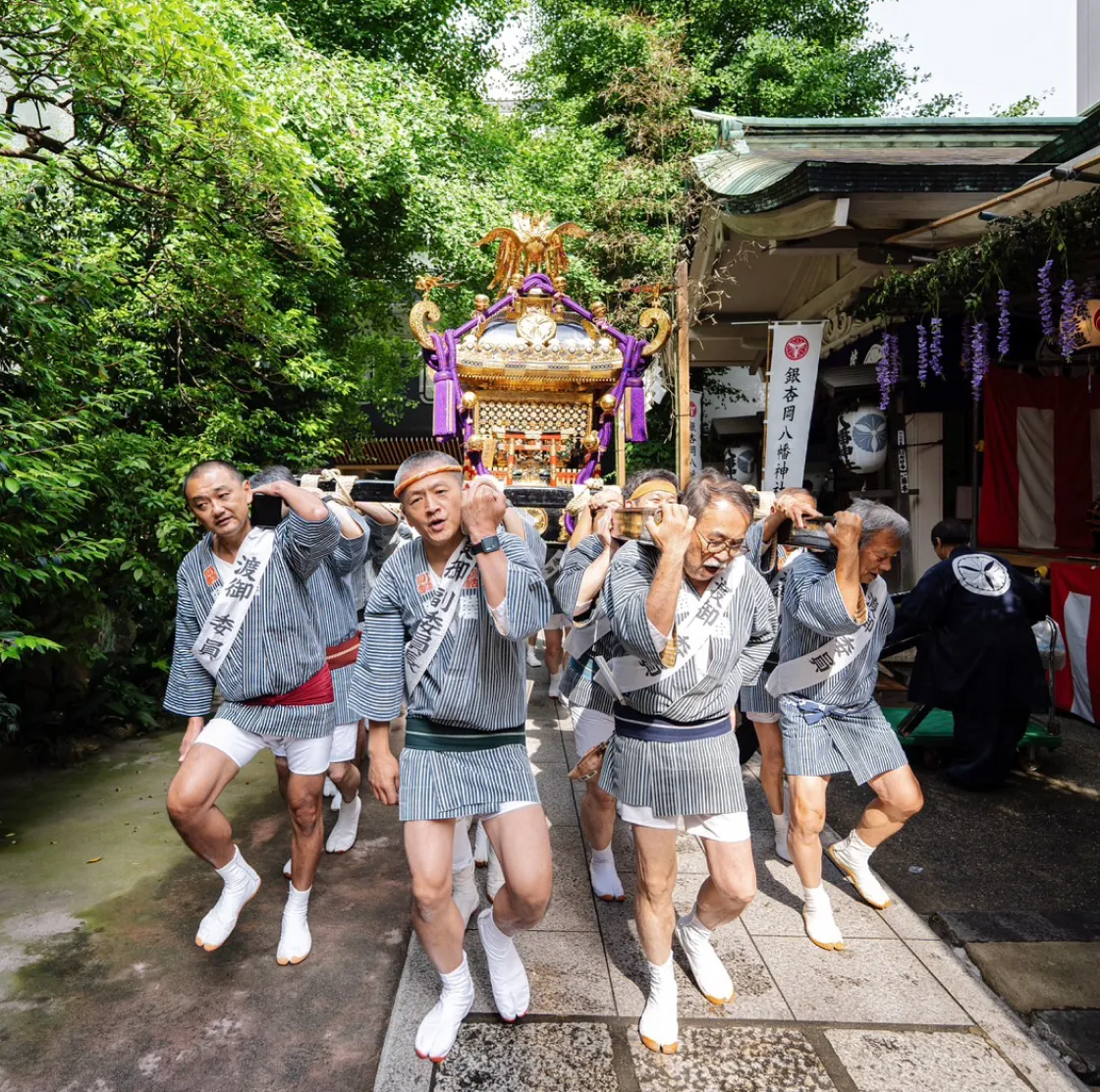 神社の参道で、法被姿の男性たちが肩に神輿を担ぎ、掛け声をかけながら行進している祭りの様子。