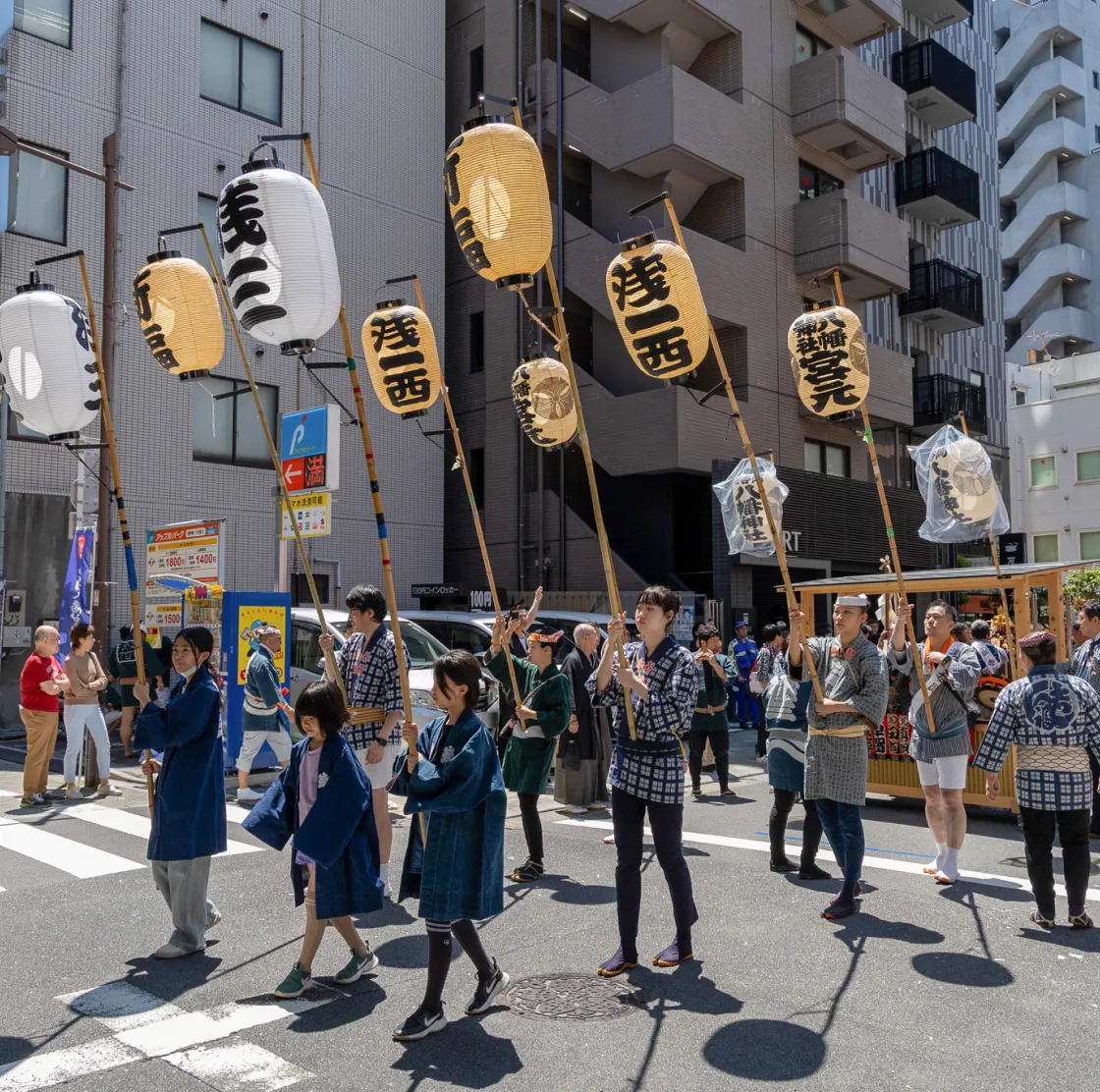 都市の交差点を行進する祭りの行列。参加者が提灯を高く掲げ、法被姿で歩く様子
