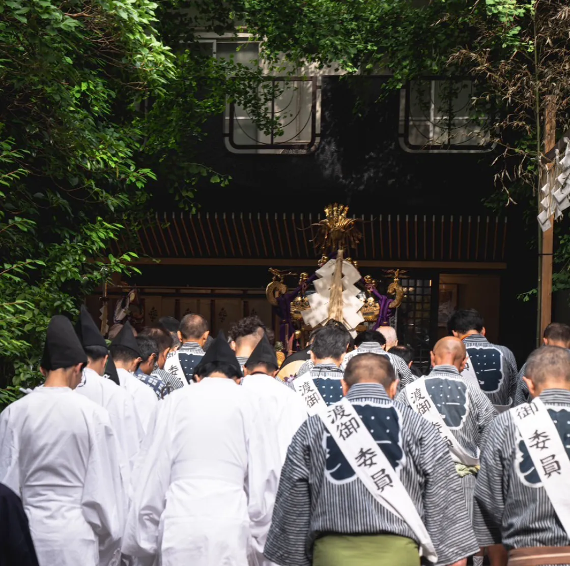 緑に囲まれた神社の参道で、法被姿の担ぎ手たちが神輿に向かって一斉に頭を下げている場面。
